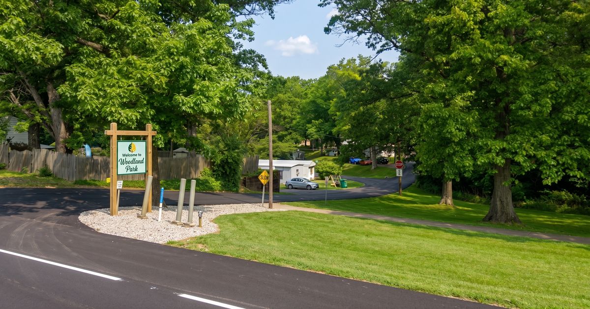 Welcome sign and paved entrance drive at a wooded mobile home community — representative of the entrance setting at Woodbine Oaks